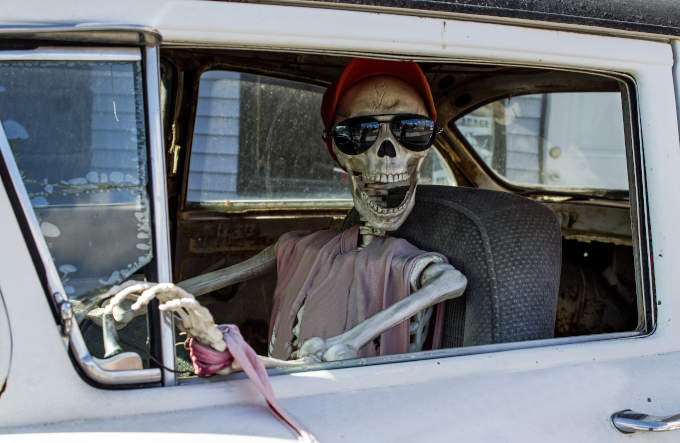 Skeleton looking at you inside an old car wearing a cap and sunglasses with its mouth open