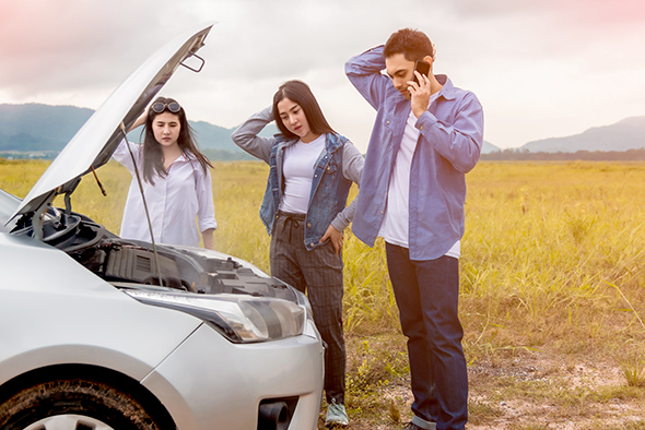 Two young women and a young man scratching their heads in front of a car with an open hood on the side of the road